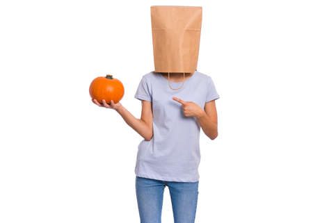 Happy halloween concept. Teen girl with paper bag over her head holds orange small fresh pumpkin, isolated on white background. Teenager cover head with bag. Child with food - raw vegetables in hands.の写真素材