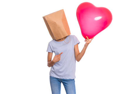 Valentines Day concept. Teen girl with paper bag over head holds red heart shaped balloon, isolated on white background. Child holding symbol of love, family, hope. Teenager cover head with bag.の写真素材