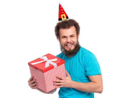 Crazy bearded Man with funny Curly Hair in little Christmas hat holding gift box. Happy guy in Santa hat, isolated on white background. Holidays concept.の写真素材