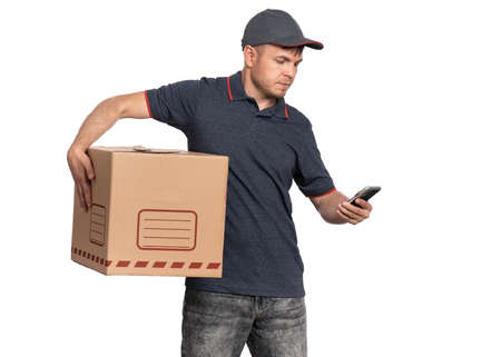 Portrait of Delivery man in cap holding cardboard box and smartphone. Handsome man checking list or searching adress on Mobile Phone. Courier isolated on white background.の写真素材