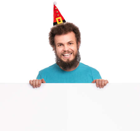 Crazy bearded Man with funny Curly Hair in Christmas hat showing empty blank signboard with copy space. Happy guy in Santa hat peeking out from behind big white banner, isolated on white background.の写真素材