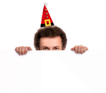 Crazy bearded Man with funny Curly Hair in Christmas hat showing empty blank signboard with copy space. Happy guy in Santa hat peeking out from behind big white banner, isolated on white background.の写真素材