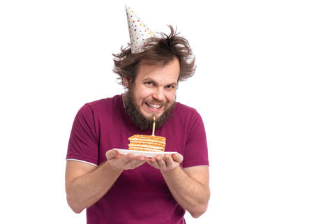 Crazy bearded Man with funny Haircut in birthday cap, isolated on white background. Guy holding plate with piece of birthday cake and burning candle. Holidays concept.の写真素材