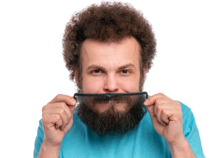 Crazy bearded Man with funny Curly Hair tries to comb his tangled and naughty hair with a small black hairbrush. Smiling guy shows mustache on his face with comb, isolated on white background.の写真素材