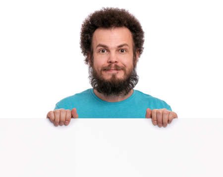Crazy bearded Man with funny Curly Hair showing empty blank signboard with copy space. Guy smiling and peeking out from behind big white banner, isolated on white background.の写真素材