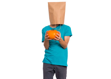 Happy halloween concept. Teen boy with paper bag over his head holds orange small fresh pumpkin, isolated on white background. Teenager cover head with bag. Child with food - raw vegetables in hands.の写真素材