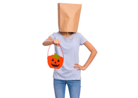 Happy halloween concept. Teen girl with paper bag over her head holds orange pumpkin bucket, isolated on white background. Portrait of teenager cover head with bag. Child waiting candy.の写真素材