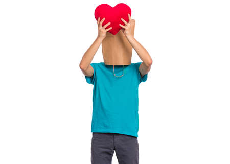 Valentines Day concept. Teen boy with paper bag over his head holds red heart, isolated on white background. Boy holding symbol of love, family, hope. Teenager cover head with bag posing in studio.の写真素材