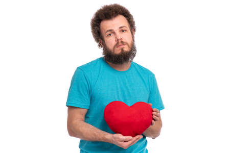 Happy Valentines Day. Crazy bearded Man with funny Curly Hair in blue T-shirt. Unhappy guy in Love, isolated on white background. Portrait of man with Red heart-shaped pillow, look at cameraの写真素材
