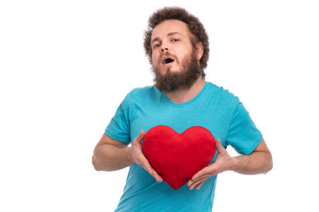 Happy Valentines Day. Crazy bearded Man with funny Curly Hair in blue T-shirt. Silly guy in Love, isolated on white background. Portrait of man with red heart shaped plush pillow, look at cameraの写真素材