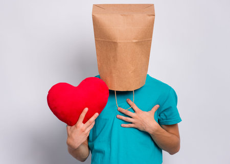 Valentines Day concept. Teen boy with paper bag over head holds red heart. Boy holding symbol of...