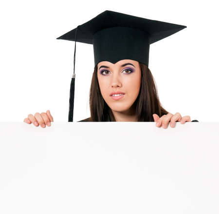Female graduate student peeking from behind a blank panel, isolated on white background. Beautiful graduate teen girl student in mantle showing blank placard board.の写真素材