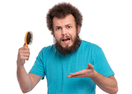 Crazy bearded Man with funny Curly Hair tries to comb his tangled and naughty hair with a small hairbrush. Surprised face of guy who holds comb, isolated on white background.の写真素材