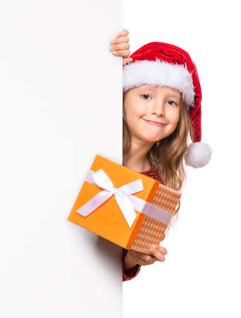 Beautiful Christmas little girl wearing red Santa hat holding banner sign. Smiling child showing white blank empty board for advertisement copyspace and gift box. Winter holidays. Happy New Year.の写真素材