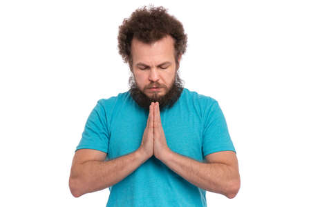 Faithful bearded handsome man stands with keeps hands together and closes eyes. Spirituality and calm atmosphere concept. Religious man praying to God with folded hands, isolated on white backgroundの写真素材