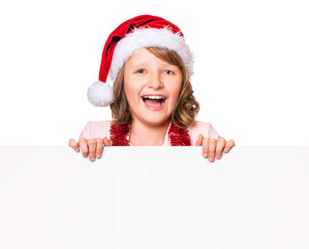 Beautiful Christmas little girl wearing red Santa hat holding banner sign. Child laughs and showing white blank empty board for advertisement copyspace. Winter holidays. Happy xmas and New Year.の写真素材