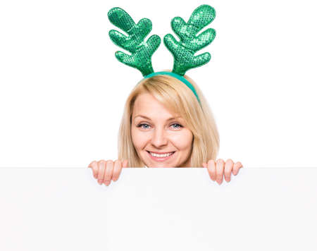 Beautiful christmas woman wearing antler deer headband holding banner sign. Smiling girl showing white blank empty board for advertisement copyspace. Winter holidays. Happy xmas and New Year.の写真素材