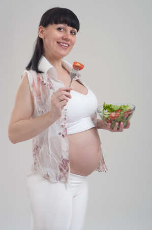 Beautiful pregnant woman holding glass bowl with fresh salad on gray background. Healthy nutrition and pregnancy. Happy pregnant woman eating vegetable salad for breakfast. Food and people concept.の写真素材