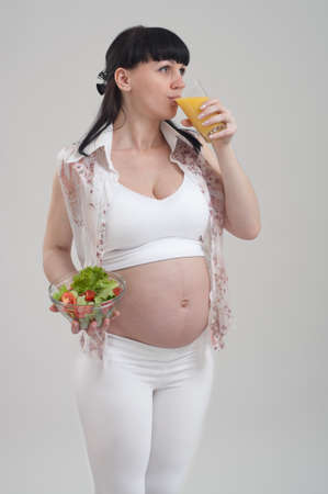 Beautiful pregnant woman holding bowl with fresh salad and drinking glass of orange juice on gray background. Healthy nutrition and pregnancy. Happy pregnant woman eating vegetable salad for breakfastの写真素材