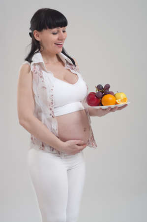 Beautiful pregnant woman holding bowl with fresh fruit on gray background. Healthy nutrition and pregnancy. Happy pregnant woman with tray of assorted fruit.の写真素材