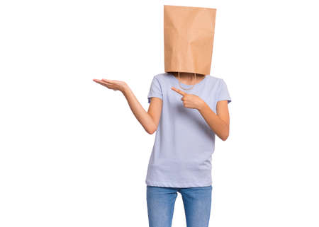 Teen girl with paper bag over head pointing finger away at copyspace, isolated on white. Teenager cover head with shopping bag pointing finger at something. Child pulling paper bag over head.の写真素材