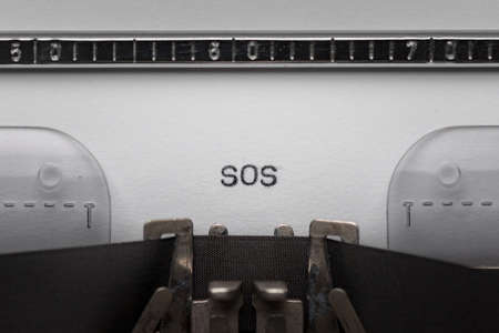 Typing text SOS on vintage manual typewriter. Close up of writes word on white paper. Typing a Morse code distress signal - The phrase save our souls.の写真素材