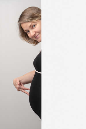 Pregnant woman peeking out of white wall. Happy Pregnant Woman touching belly with hand fingers. Baby expectation. Playing on baby bump.の写真素材