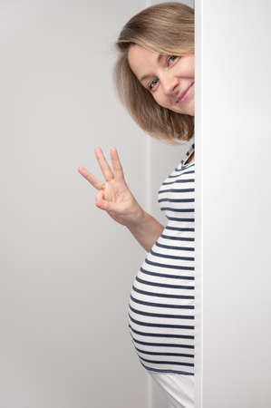 Pregnant woman peeking out of white wall and showing three fingers sign. Happy Pregnant Woman looking at camera and smiling. Baby expectation.の写真素材