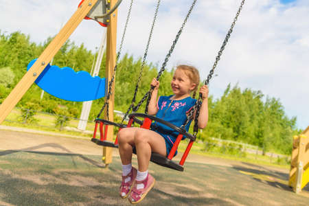 Happy child girl on swing on warm and sunny day outdoors. Little kid playing on nature walk in playground in park, cute blond girl swinging high, active summer leisure for kids, joyful holidaysの写真素材