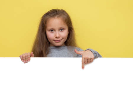 Place for your advertisement. Baby girl pointing to blank white billboard on yellow background, copy space. Emotionsの写真素材