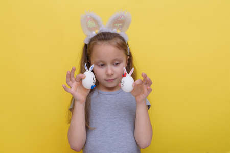 Cute little child wearing bunny ears on Easter day. Girl holding a painted egg on light yellow background.Studio photography.の写真素材