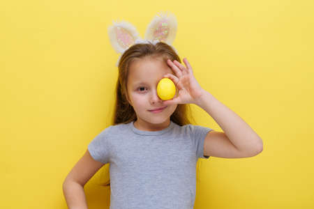 Cute little child wearing bunny ears on Easter day. Girl holding a painted egg on light yellow background.Studio photography.の写真素材