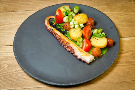 Octopus tentacles with baked potatoes and vegetables .On black plate. On a rustic wooden backgroundの写真素材