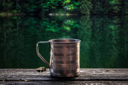 Steel cup of hot tea on wooden surface. Forest lake in background.の写真素材