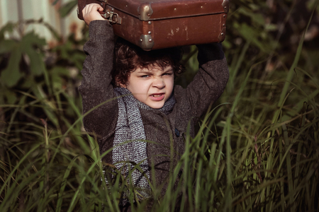A little boy in a coat sneaks through the tall grass, with a suitcase on his head. Little traveler. Tourism. Vintage. Escape from home.の写真素材