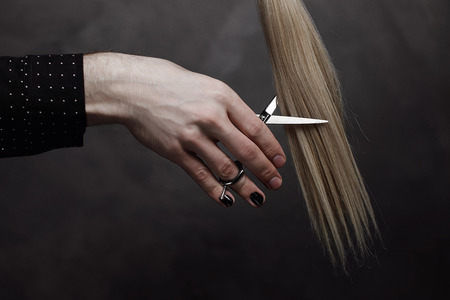 The male hand with manicure holds scissors and a lock of hair on a gray background. Nails are painted black color. Close up.の写真素材
