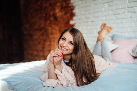 A beautiful young brunette in a light shirt laughs while lying in bed in her modern bedroom. Cute girl resting at home in the winter.の写真素材