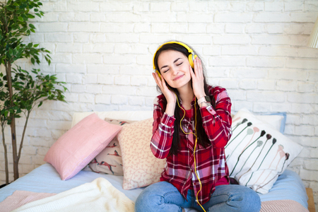 Happy pretty woman in earphones listening to music and singing while sitting on bed and holding mobile phoneの写真素材