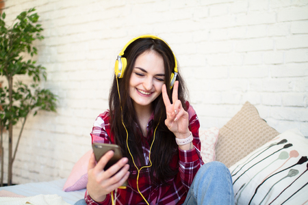 Happy pretty woman in earphones listening to music and singing while sitting on bed and holding mobile phoneの写真素材