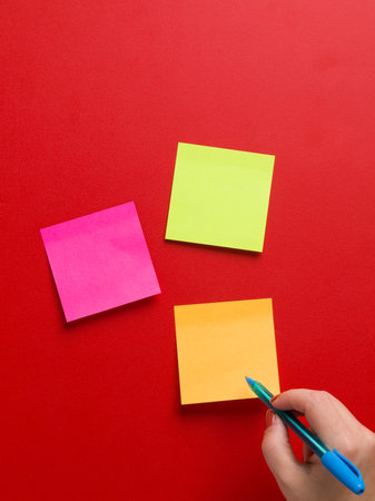Top view of three colored reminder stickers and a female hand holding a blue pen in a red...