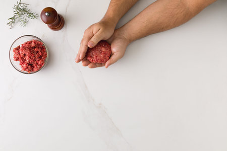 Top view of male hand molding a hamburger in a marble table, a glass bowl with meat, wooden pepper...