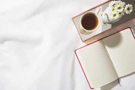 Top view of morning scene in voile fabric background with a red notebook, mug of tea and a vase of...