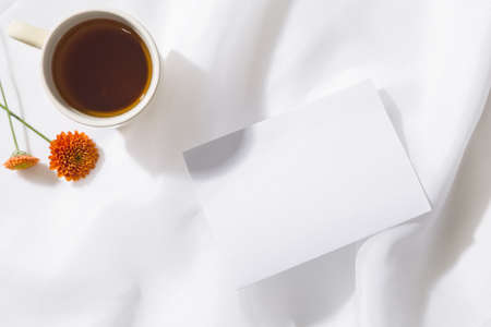 Top view of voile fabric background with a mug of tea, two orange flowers and a piece of white paper with space for text. Horizontalの写真素材