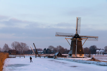 A ice skater is on his way  home at sunset, passing an old mill in winter in the Netherlandsの写真素材