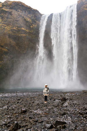 Beautiful female tourist dwarfed in front of the mighty Skogafoss waterfall in Icelandの写真素材