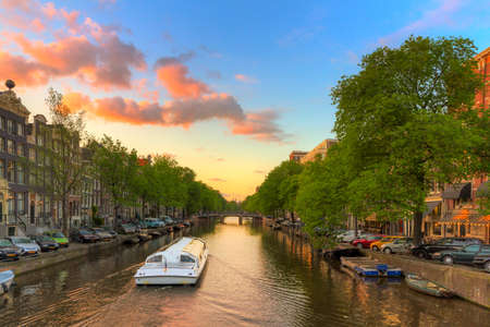 Tourist canal boat tour at sunset in one of the beautiful canals of Amsterdam, The Netherlandsの写真素材