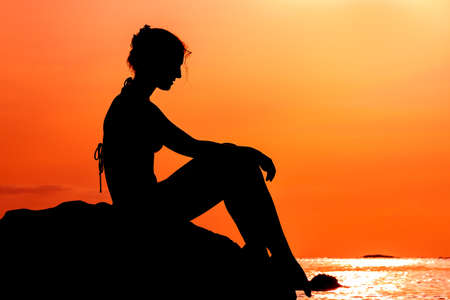 Silhouette of a beautiful woman sitting on a rock at sunset looking at the pacific ocean in summer in Puerto Ricoの写真素材