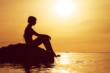 Silhouette of a beautiful woman sitting on a rock at sunset looking at the pacific ocean in summer in Puerto Ricoの写真素材