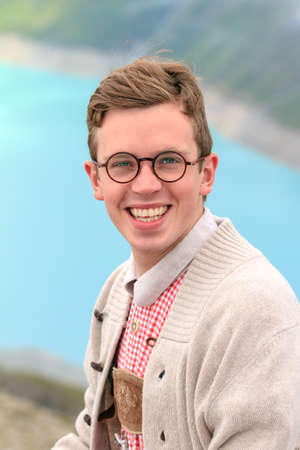 Beautiful portrait of a handsome young man in traditional clothing with lederhosen and checkered shirt in summer in the alps near Grimentz, Switzerland, with blue lake Lac de Moiry in the backgroundの写真素材