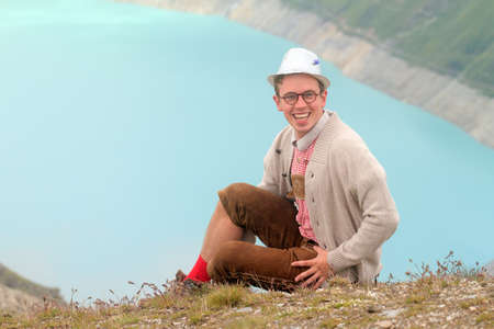 Beautiful portrait of a handsome young man in traditional clothing with lederhosen and checkered shirt in summer in the alps near Grimentz, Switzerland, with blue lake Lac de Moiry in the backgroundの写真素材